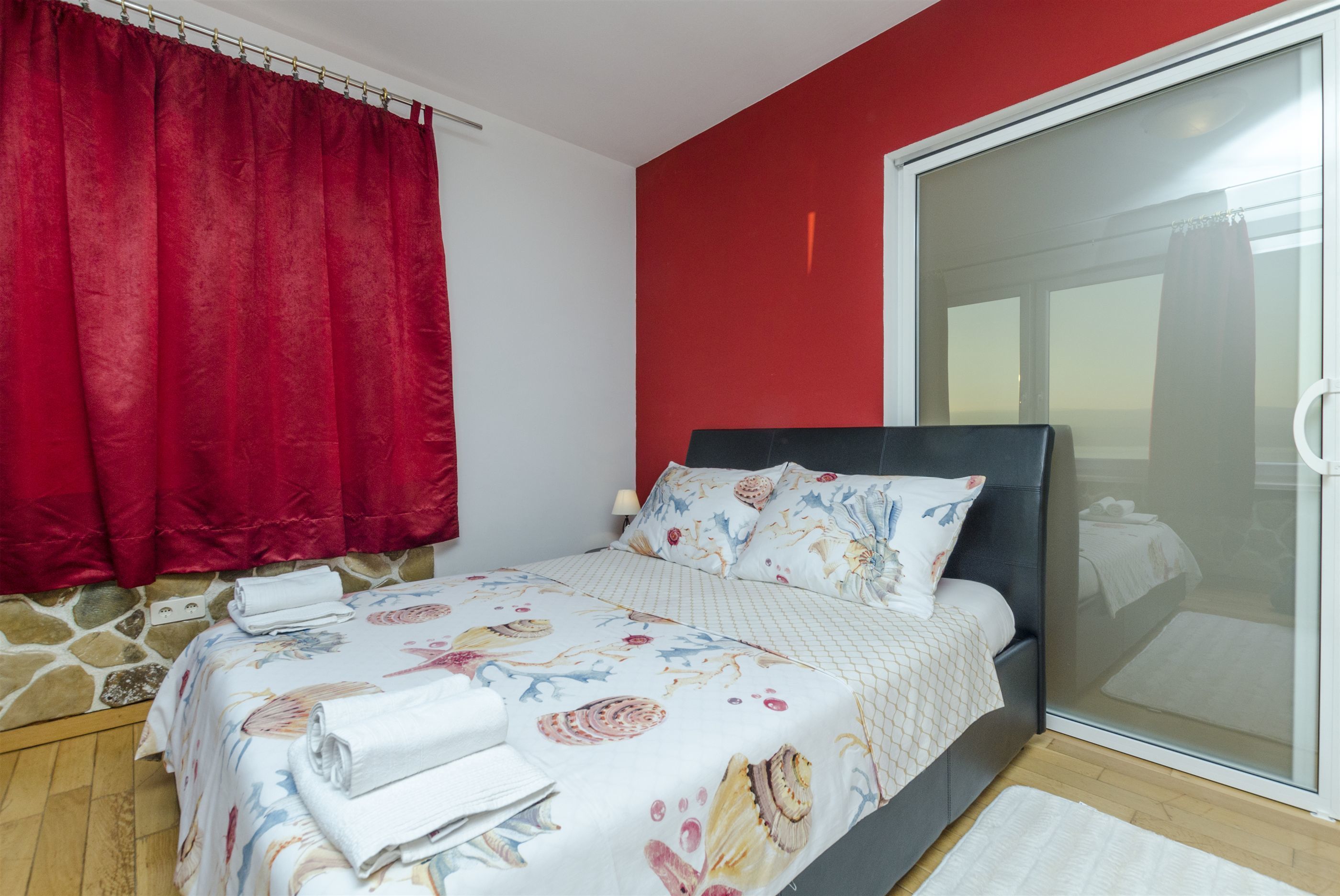 A bedroom with a bed, red curtains, a stone wall, and a sliding glass door.