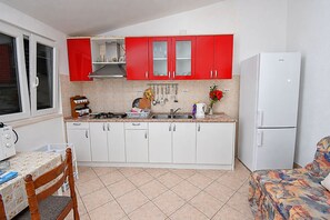 A compact kitchen with red and white cabinets, a white refrigerator, and a dining area with a table and chairs. - Apartments Srdan (Rogoznica)