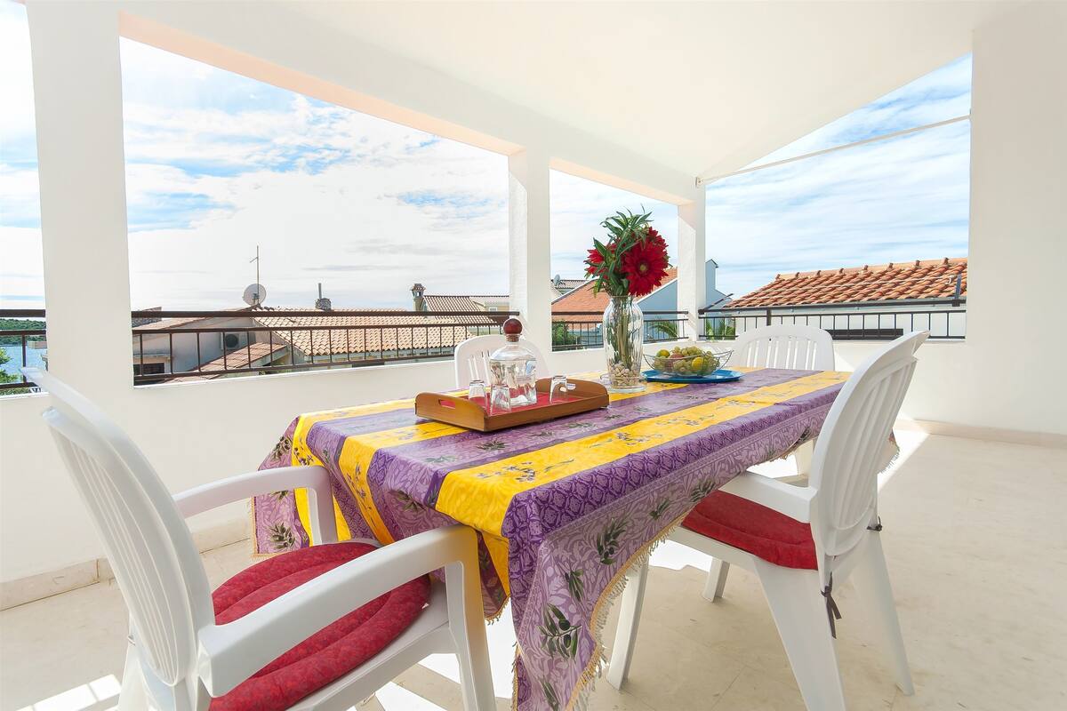 A balcony with a table set for dining, white chairs, a vase with flowers, and a view of buildings and a body of water.
