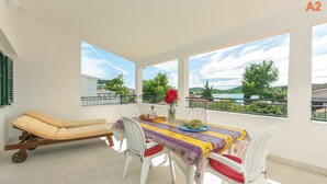 A room with a table, chairs, a sun lounger, and large windows with a view of trees and water. - Apartments Srdan (Rogoznica)
