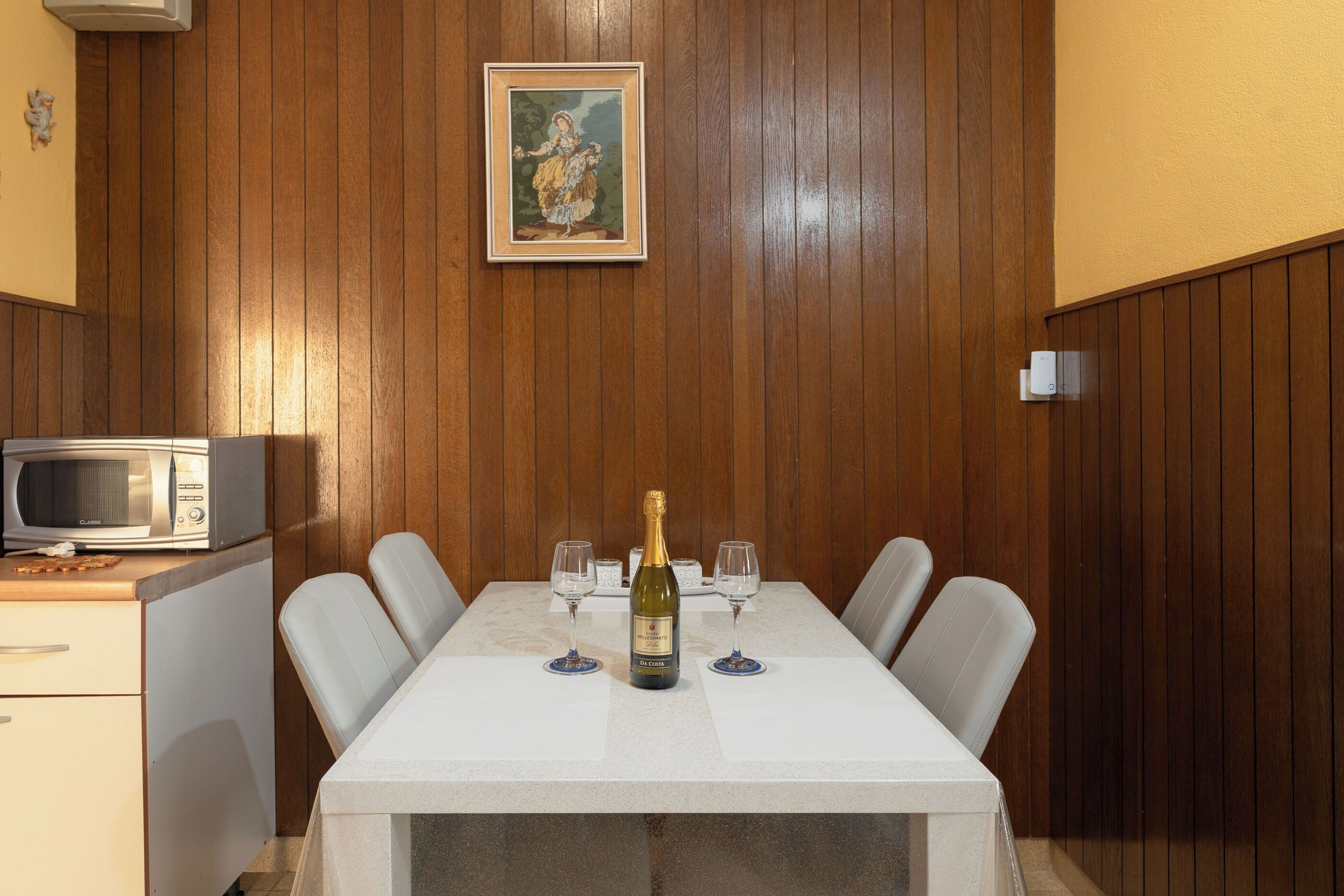 A dining table with a bottle of champagne, two wine glasses, and a microwave on a wooden-paneled wall.