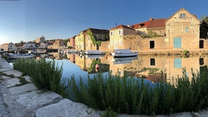 A waterfront scene with stone buildings, a calm water body, and boats docked along the shore. - House Tia (Vrboska)