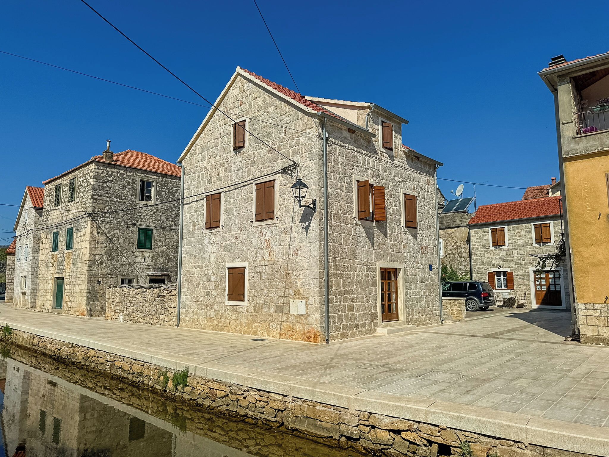 A stone building with a tiled roof, a paved street, and a body of water reflecting the buildings.