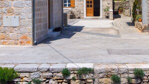 A stone building with a wooden door and windows, a stone wall with plants, and a clear blue sky. - House Tia (Vrboska)