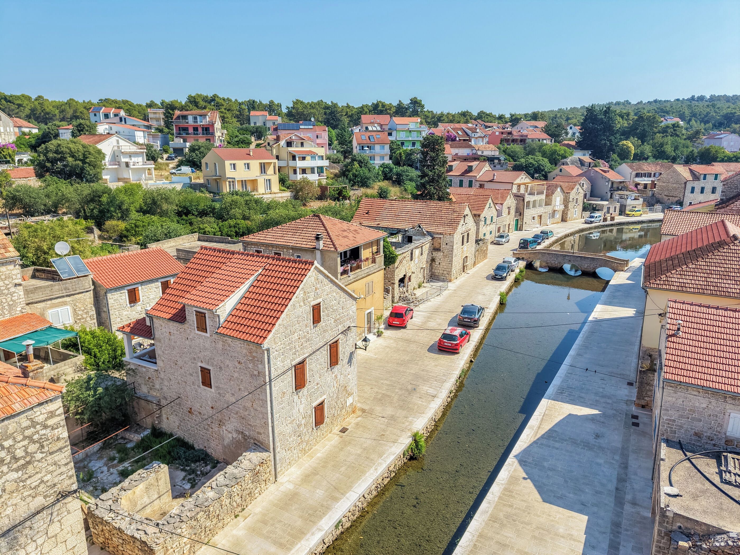 A narrow street with buildings on either side, a canal, and parked cars.