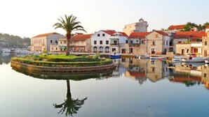 A waterfront town with historic buildings, a central palm tree, and calm water reflecting the architecture. - House Tia (Vrboska)