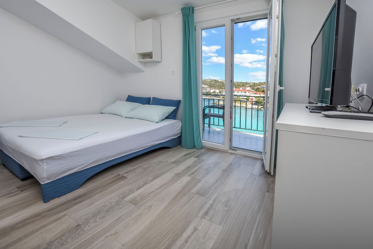 A bedroom with a bed, a desk with a computer, and a balcony with a view of the sea.
