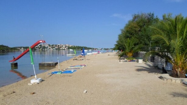 A sandy beach with a red slide, palm trees, and a calm body of water in the background. - Apartments Miodrag (Stupin Celine)