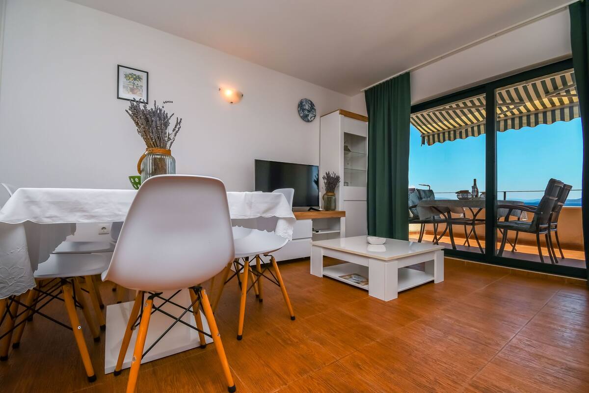 A modern living room with a white sofa, a dining table with chairs, a coffee table, a TV, and a bookshelf. There is a balcony with a view of the sky and a table with chairs.