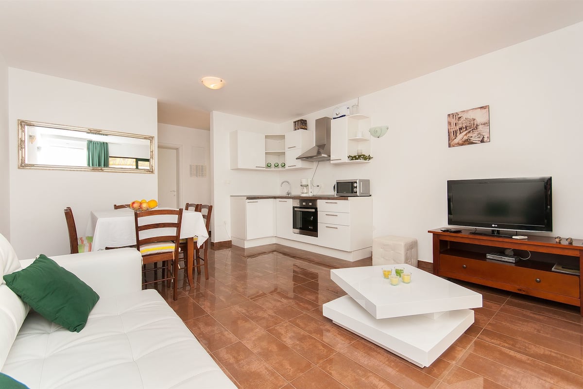 A modern living room with a sofa, coffee table, and a TV. There's a kitchen area with a microwave and oven.