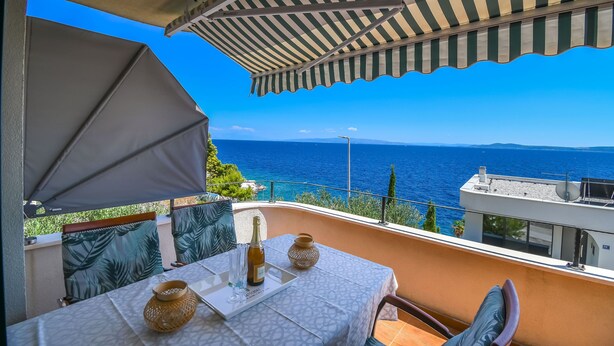 A balcony with a table set for two, a bottle of wine, and a vase. The balcony overlooks a scenic view of the sea. - Apartments Sanja (Okrug Gornji)