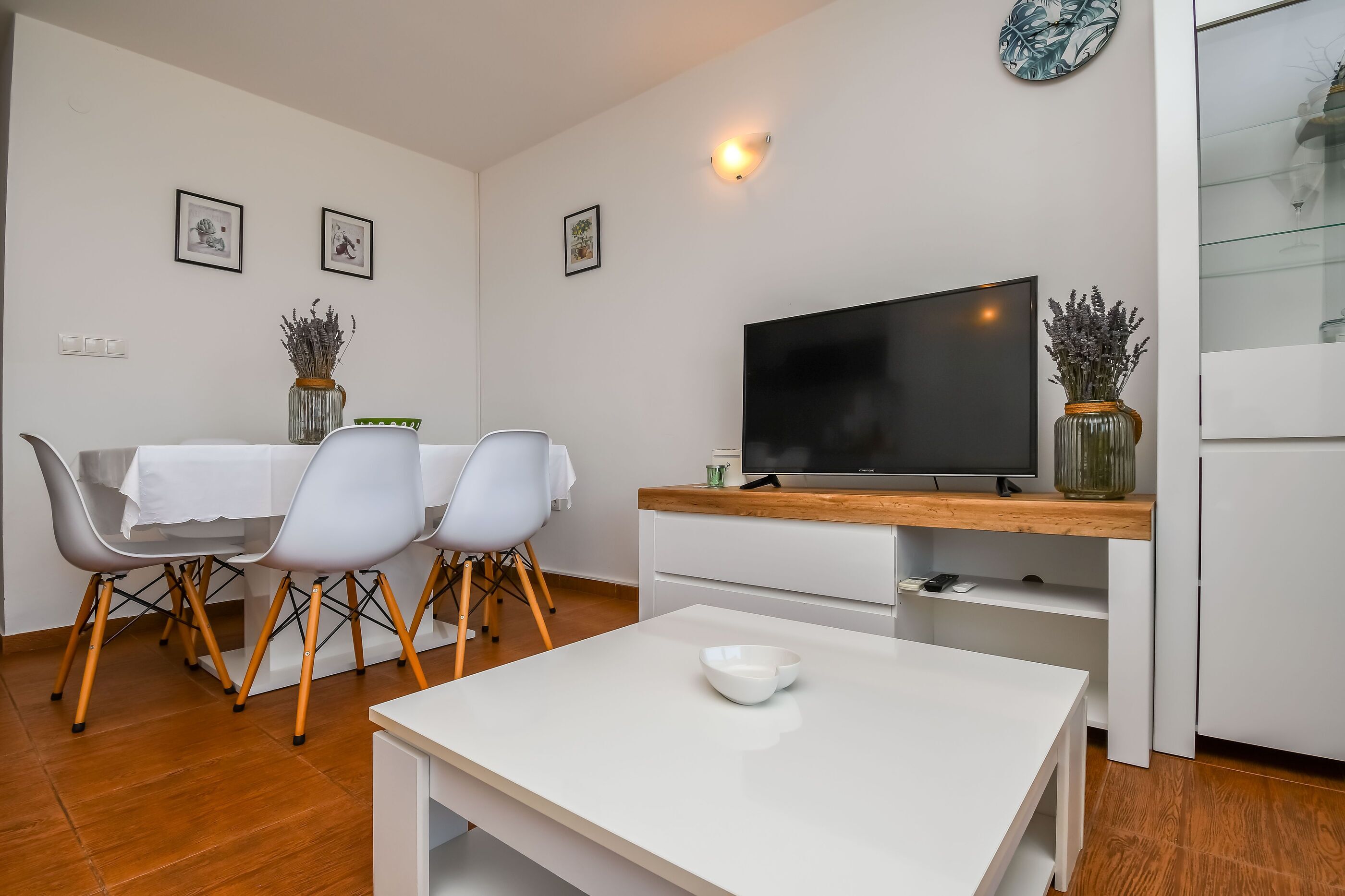 Una sala de estar moderna con comedor, muebles blancos, un televisor de pantalla plana y jarrones decorativos.