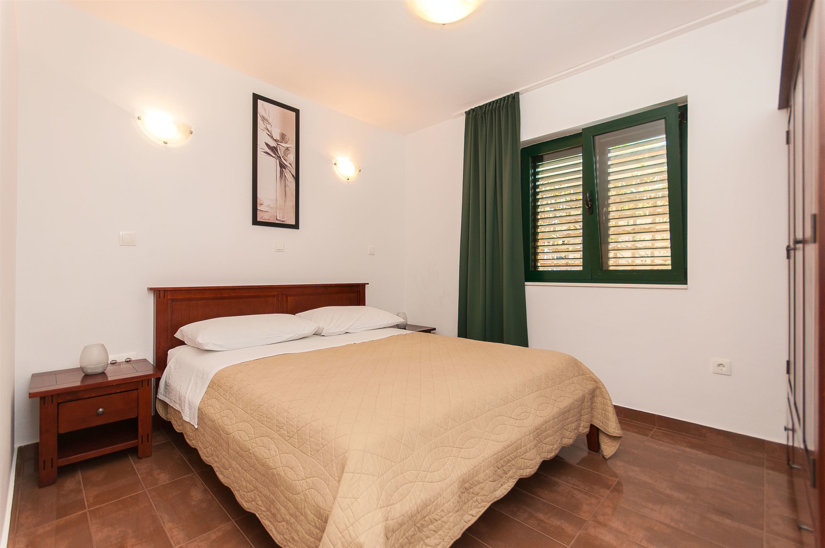 Une chambre à coucher comprenant un lit, une table de chevet, une lampe fixée au mur, une fenêtre avec des stores et un tableau encadré.