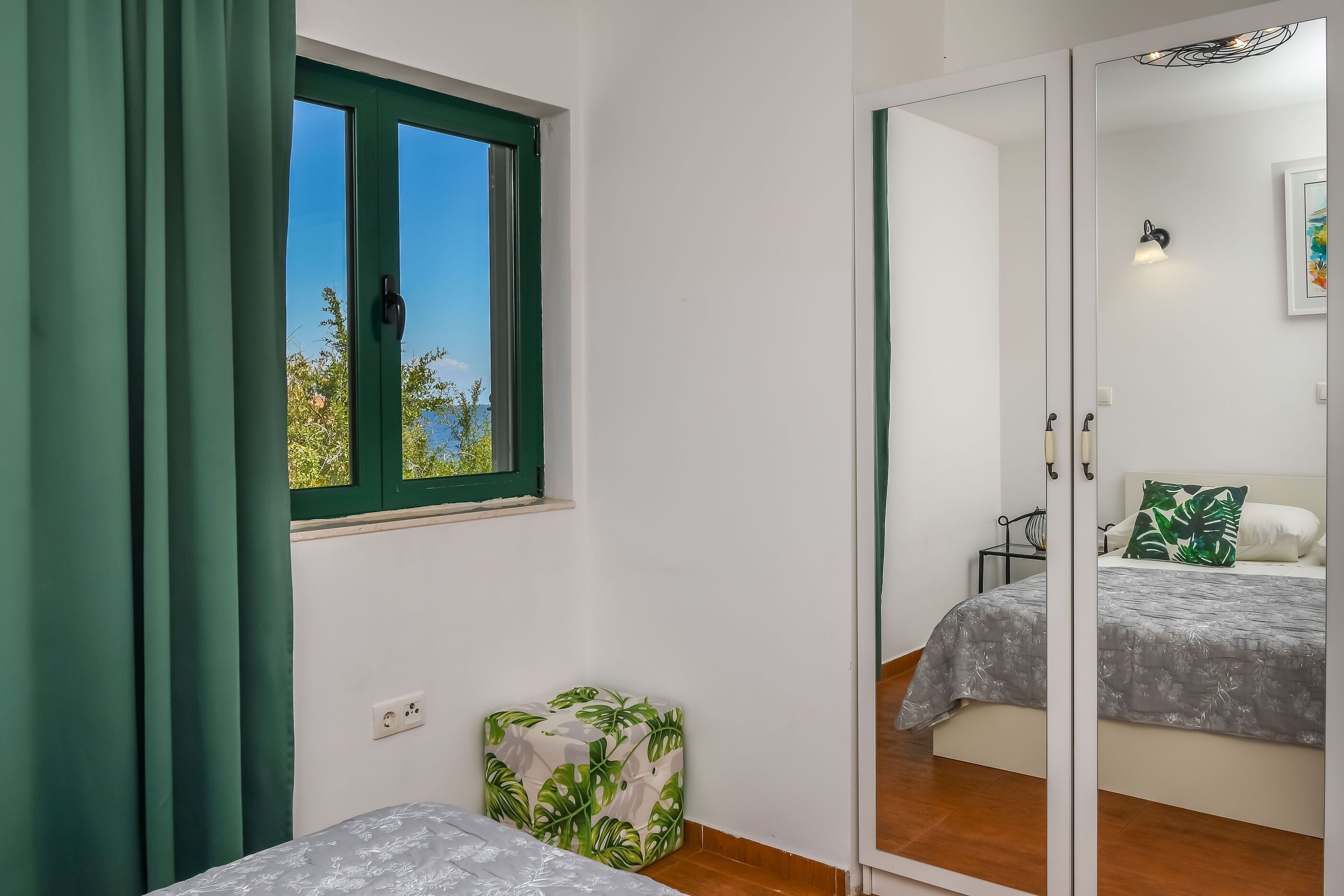 A bedroom with a bed, a small stool, a window with curtains, and a mirror.