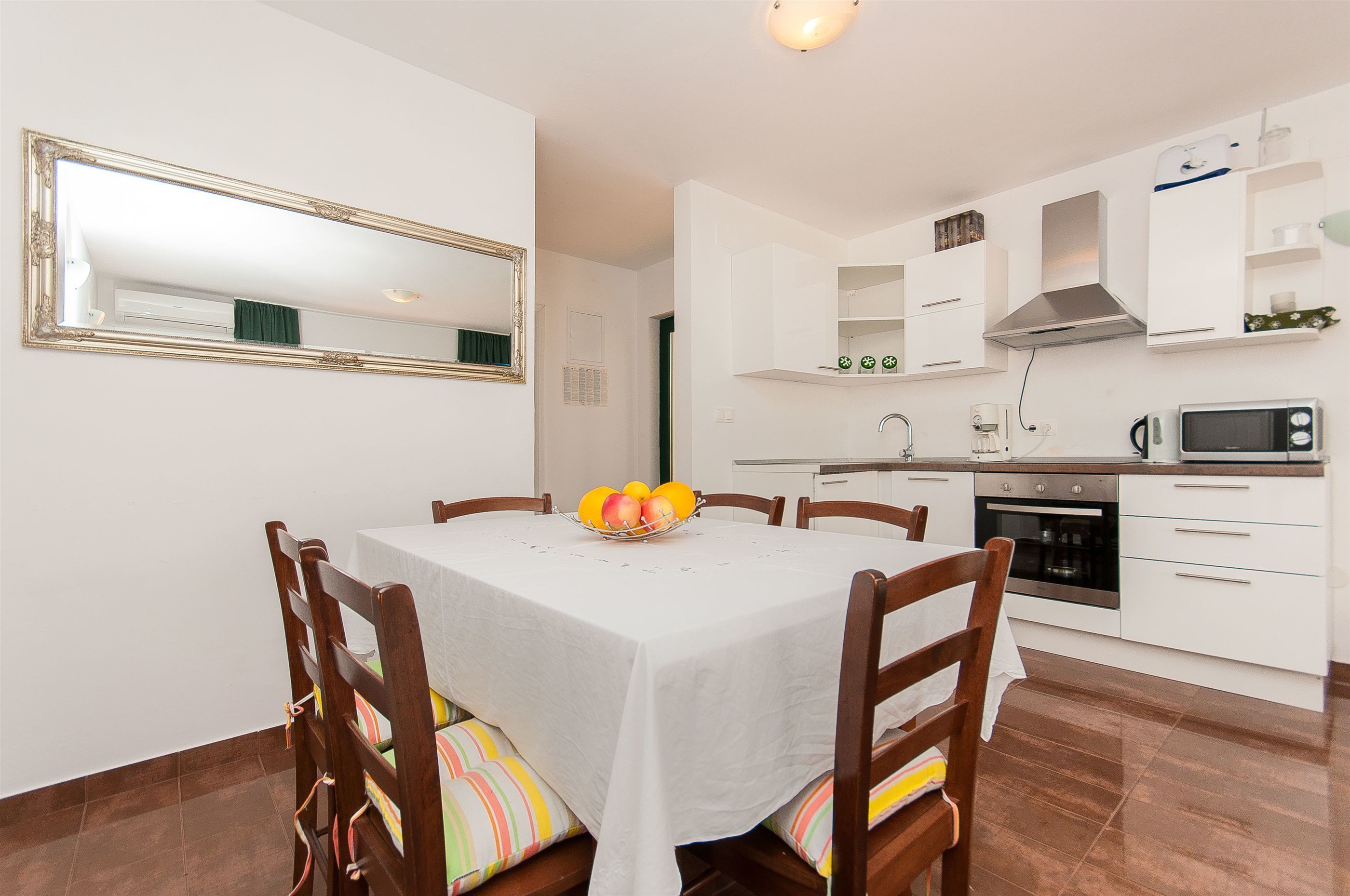 Een eetgedeelte met een tafel voor vier personen, een fruitschaal, en een keuken met witte kasten en apparatuur.