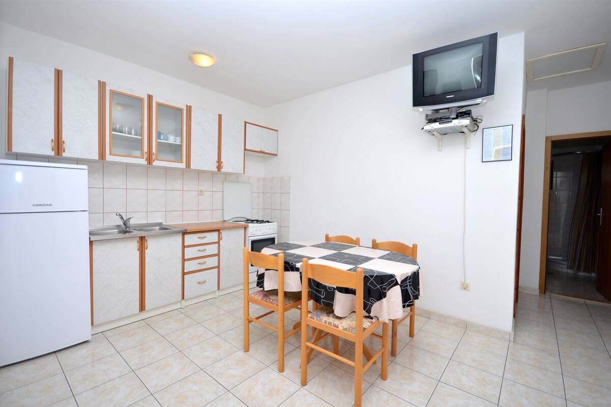 A compact kitchen with a dining table, wooden cabinets, a built-in oven, and a television mounted on the wall.