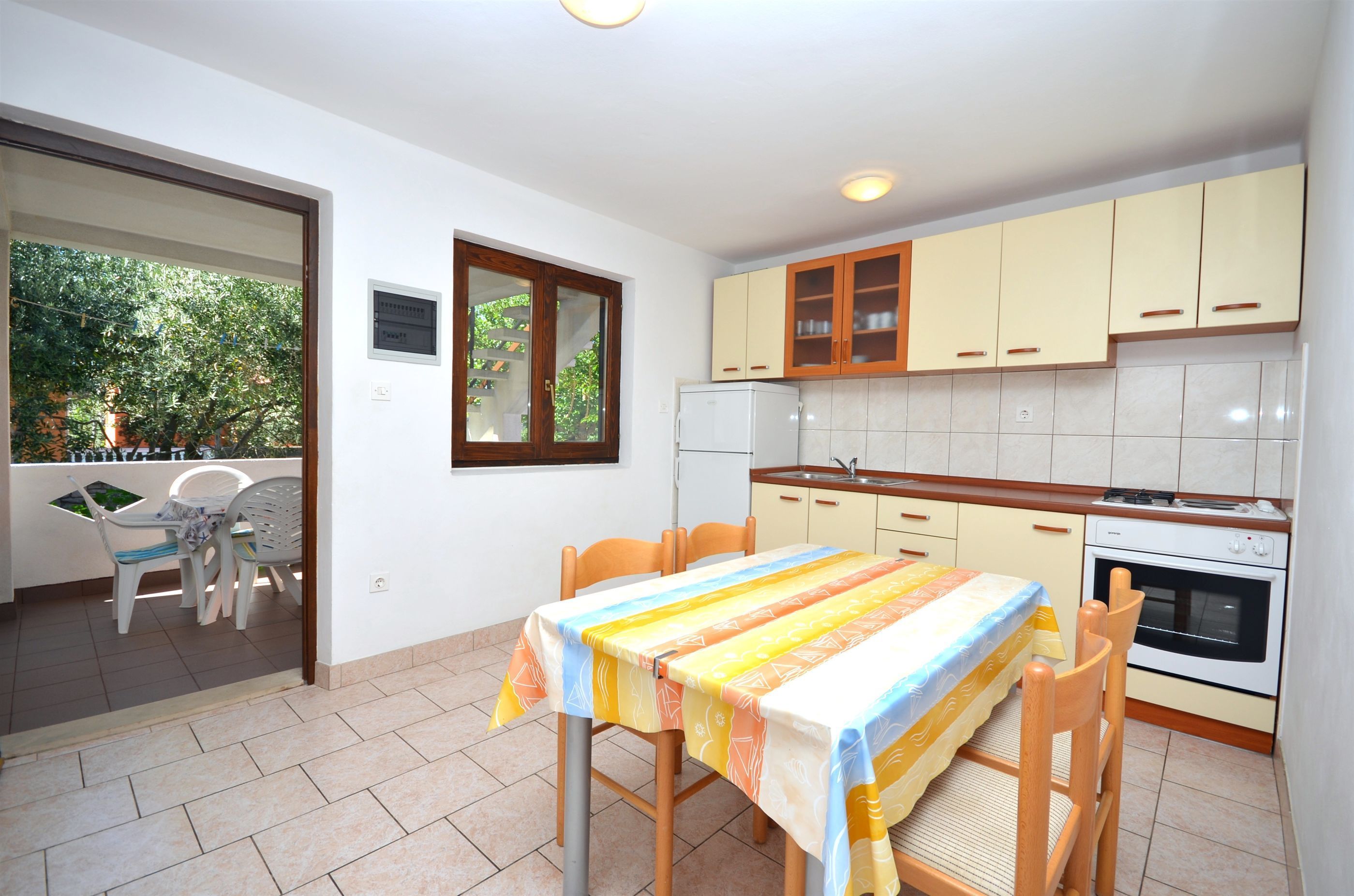 A compact kitchen with wooden cabinets, a dining table with chairs, and a window overlooking greenery.