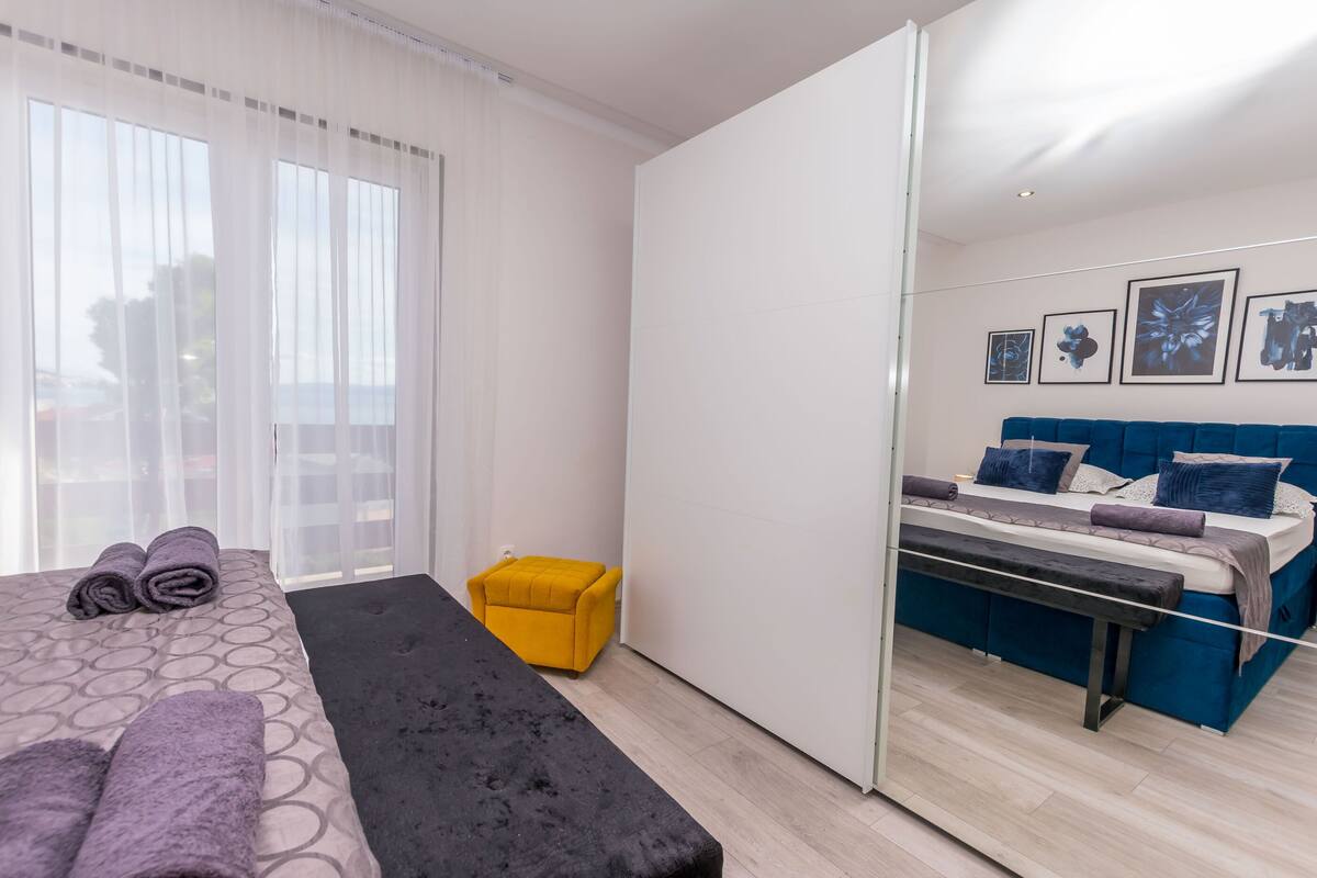 A modern bedroom with a bed, a bench, a yellow ottoman, and a large mirror.
