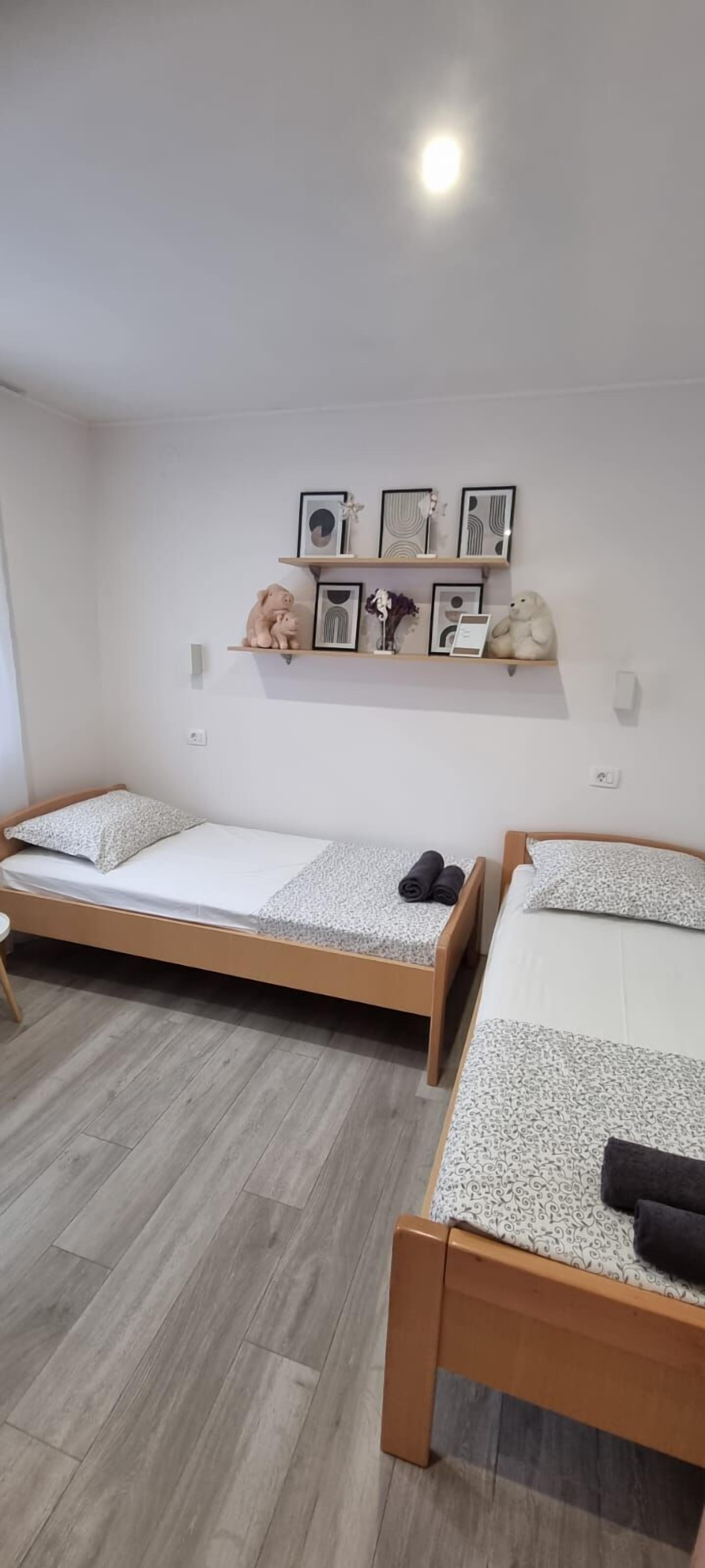 A room with two wooden beds, a white wall with a shelf displaying framed artwork and decorative items, and a wooden floor.