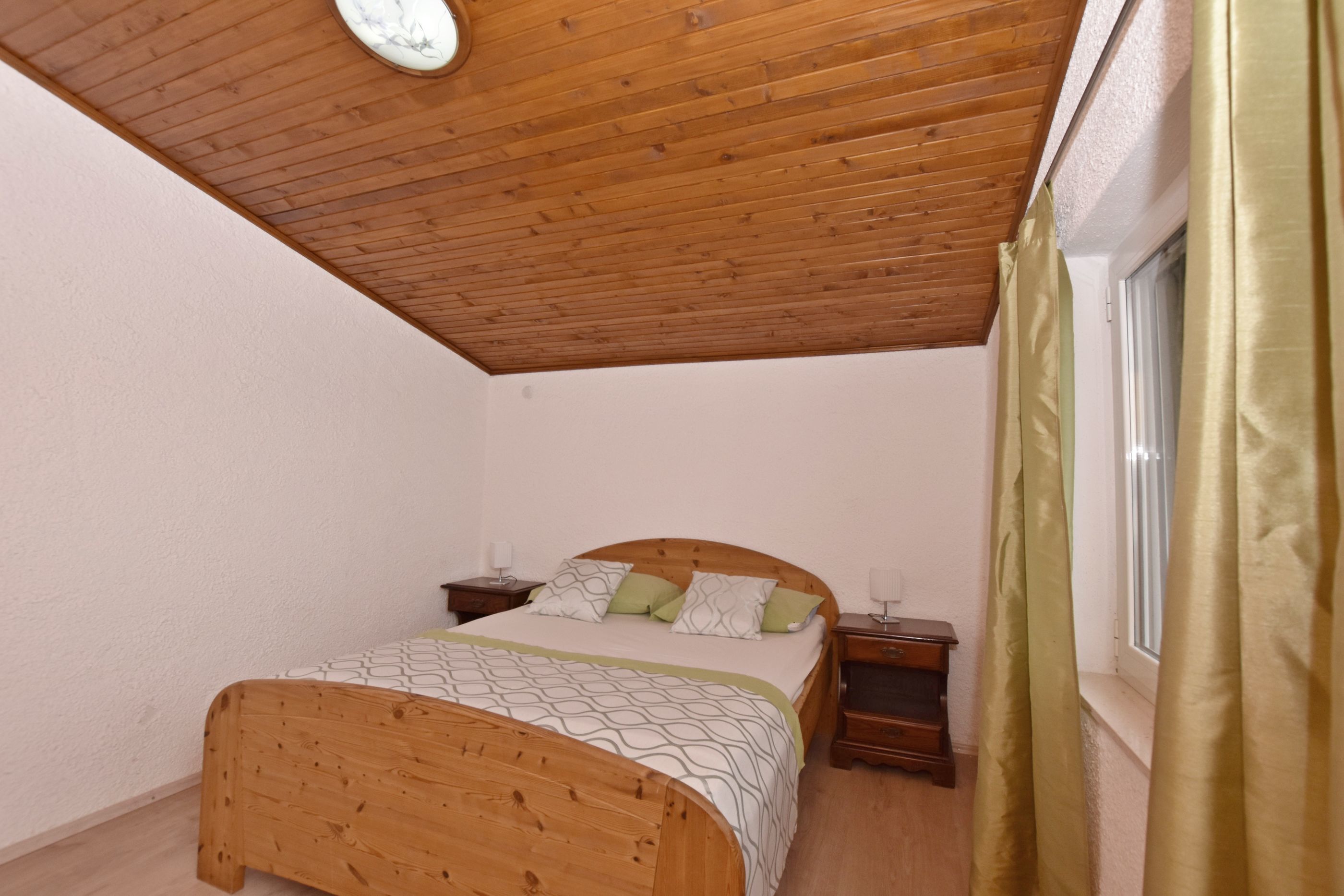 A bedroom with a wooden ceiling, a bed with a patterned bedspread, two bedside tables, and a window with curtains.