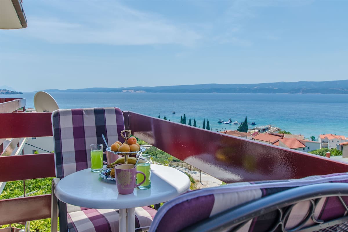 A balcony with a table set for two, overlooking a coastal view with boats and buildings.