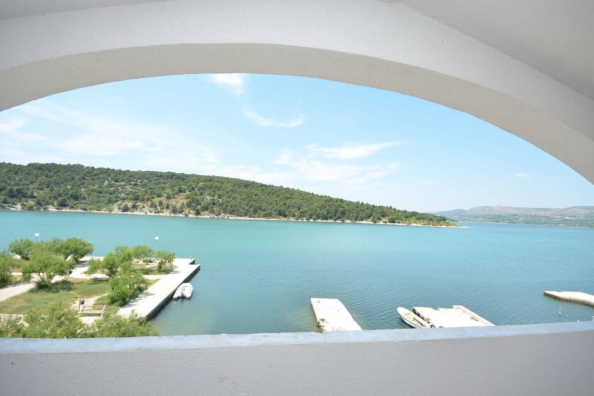 Vista su un lago dal balcone, cielo sereno, barche ormeggiate e una panchina.