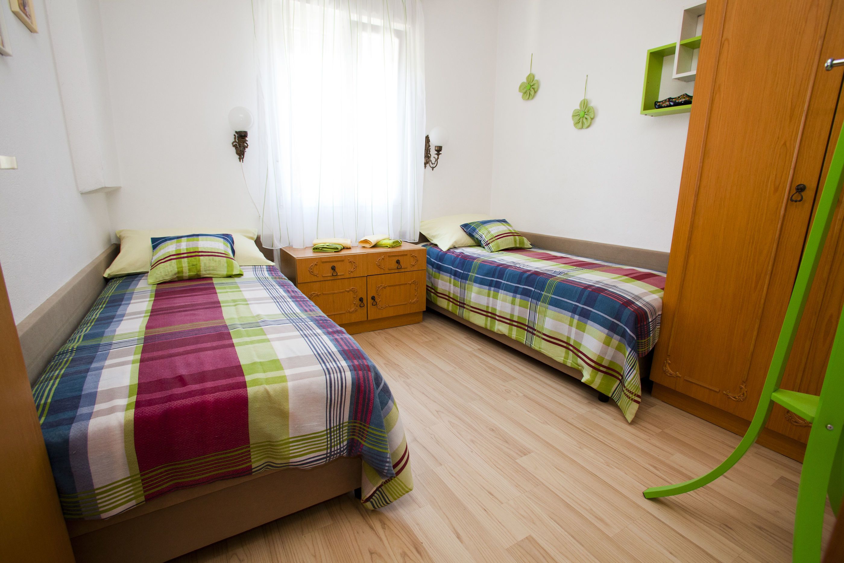 A bedroom with two beds, a wooden dresser, a green chair, and a window with sheer curtains.