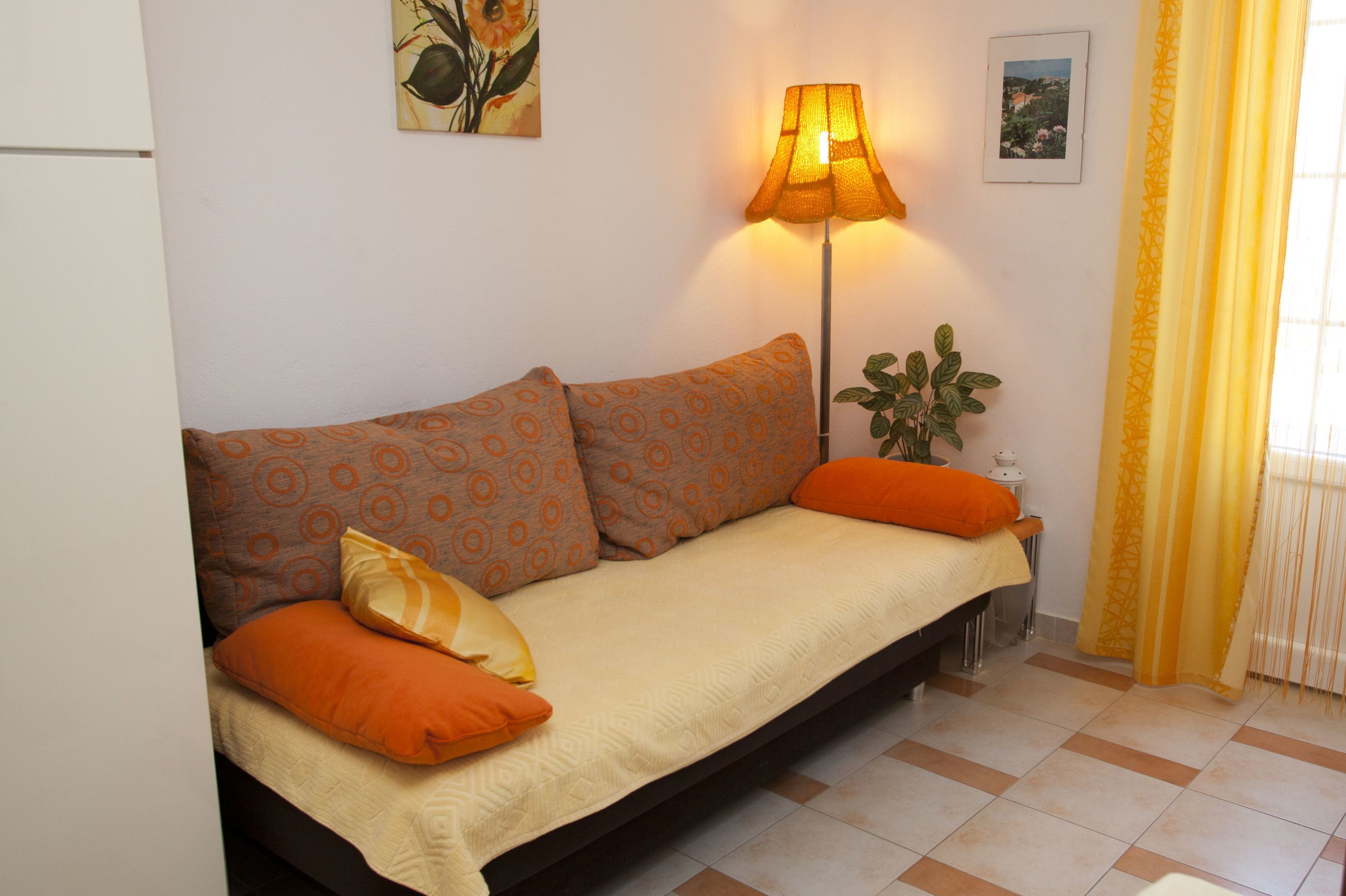 Un sofá con cojines estampados, una lámpara de pie y una planta en una habitación con suelo de baldosas y una ventana con cortinas amarillas.