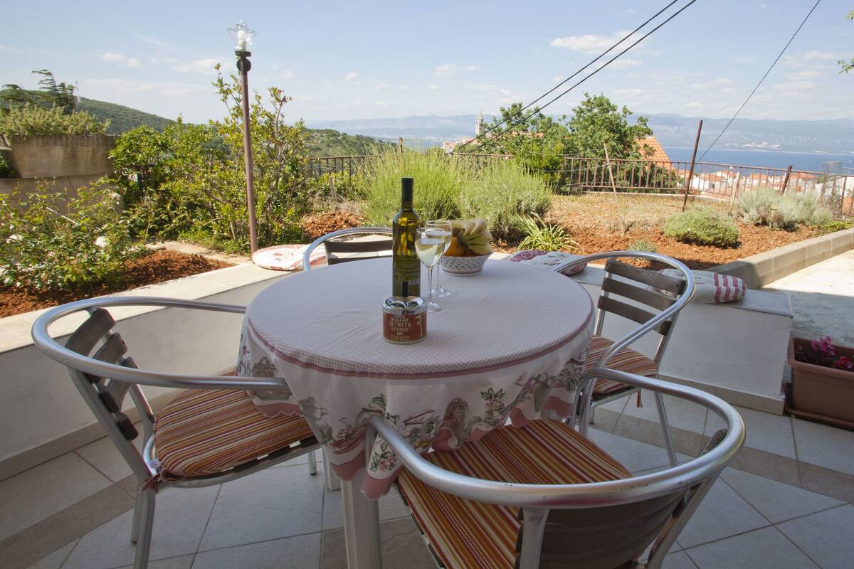 Une table ronde recouverte d’une nappe à motifs floraux, entourée de chaises en métal avec des coussins à rayures, est placée sur une terrasse carrelée d’où l’on peut admirer des collines et la mer.