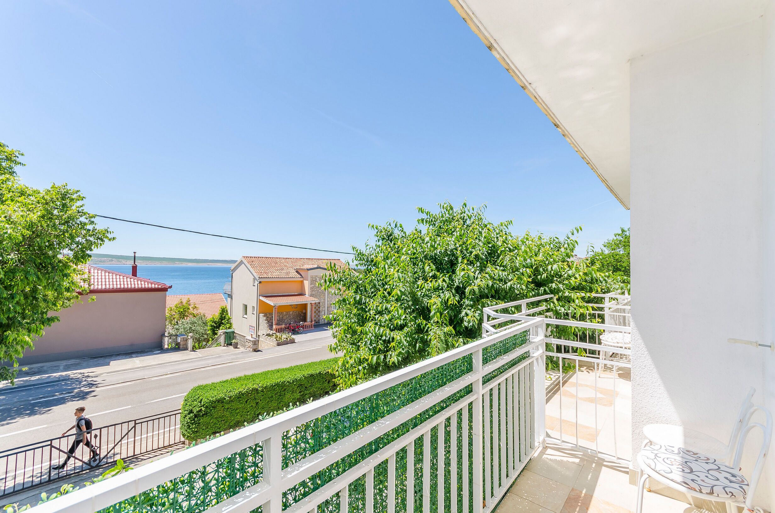 A balcony with a white railing overlooking a street, a clear blue sky, and a body of water in the distance.