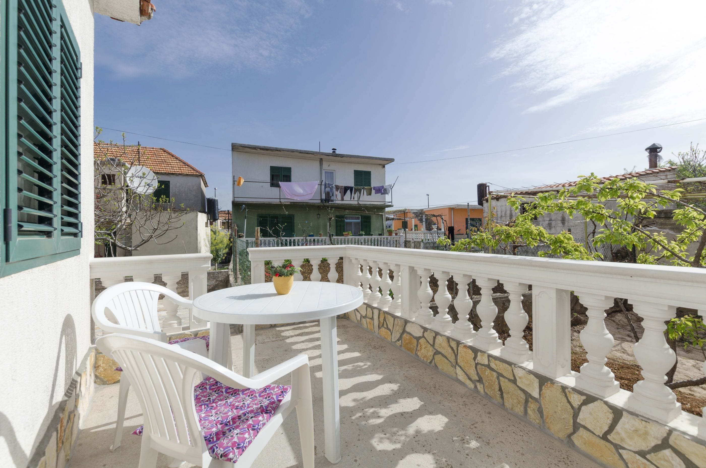A balcony with a white table and chairs, a stone balustrade, and a view of a building with a tiled roof.