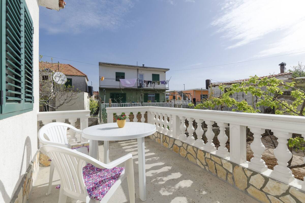 A balcony with a white table and chairs, a stone balustrade, and a view of a building with a tiled roof.
