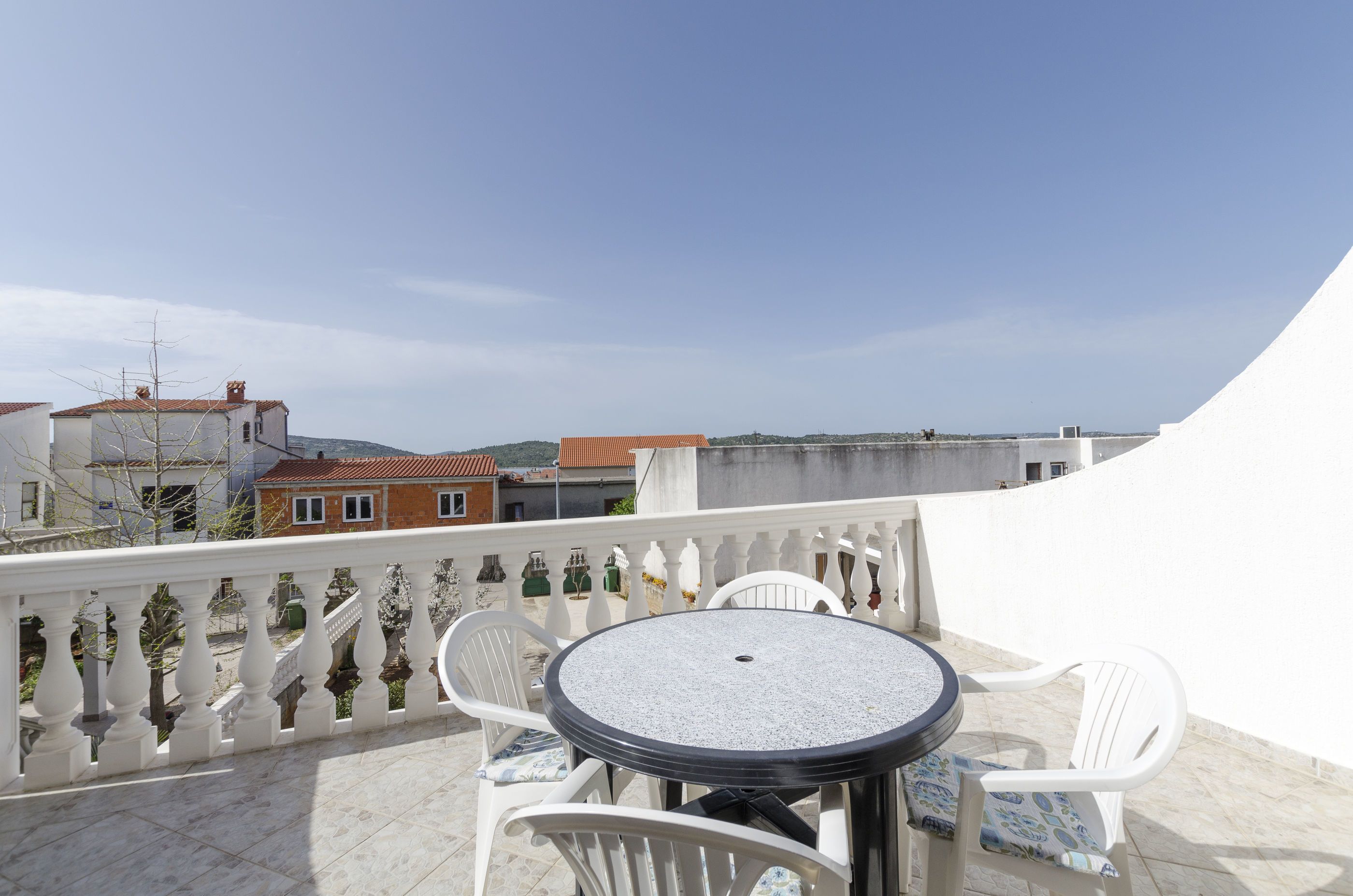 A balcony with a round table and white chairs, overlooking a cityscape with buildings and a clear sky.