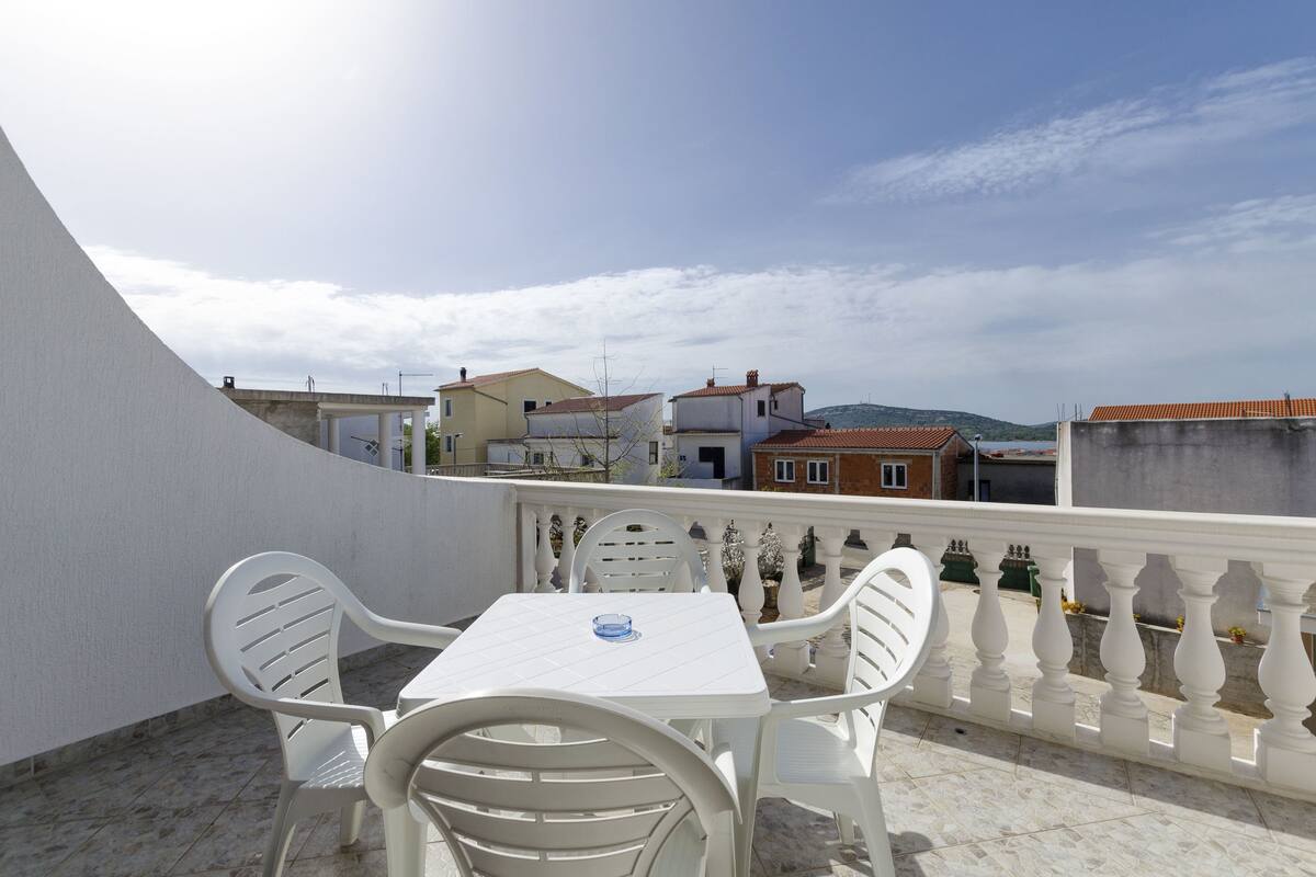A balcony with a white table and chairs, overlooking a residential area with houses and a hilly landscape.