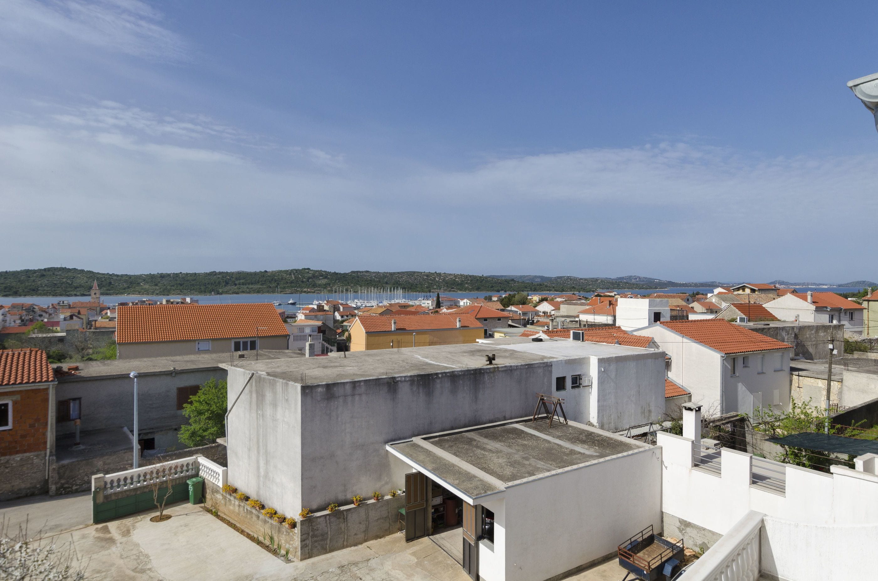 A residential area with multiple buildings, some with red-tiled roofs, and a clear view of the sea in the background.