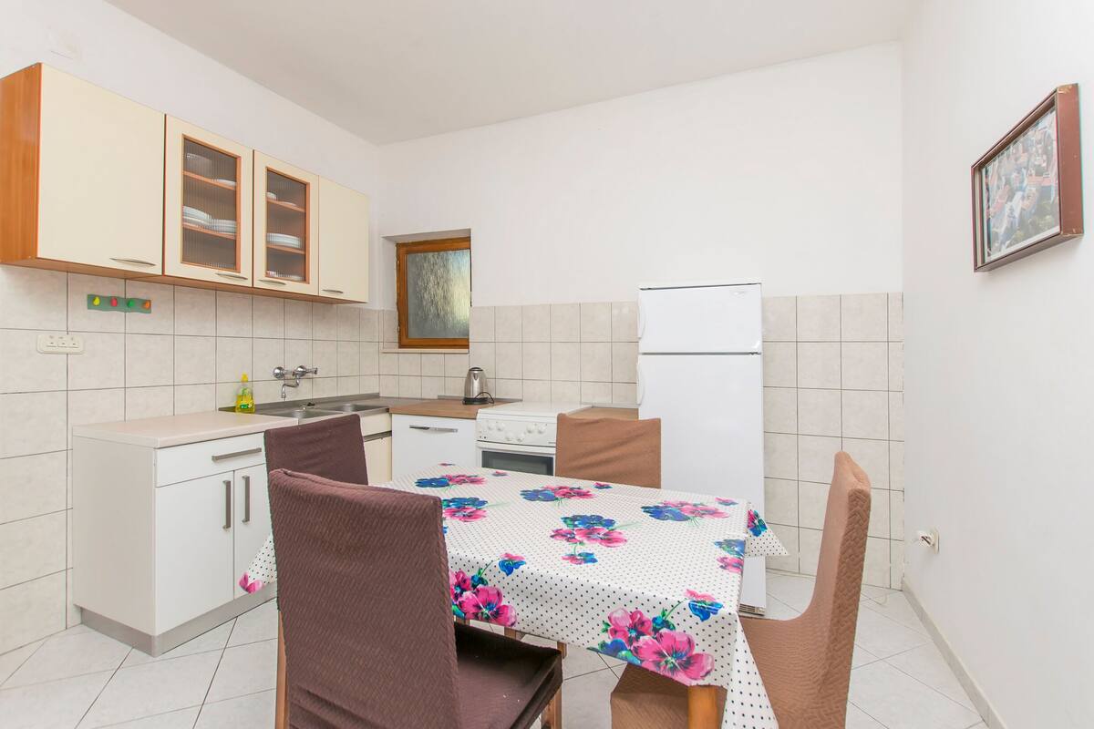 Une cuisine de taille réduite avec des meubles blancs, une table à manger recouverte d’une nappe à motifs floraux et un réfrigérateur.