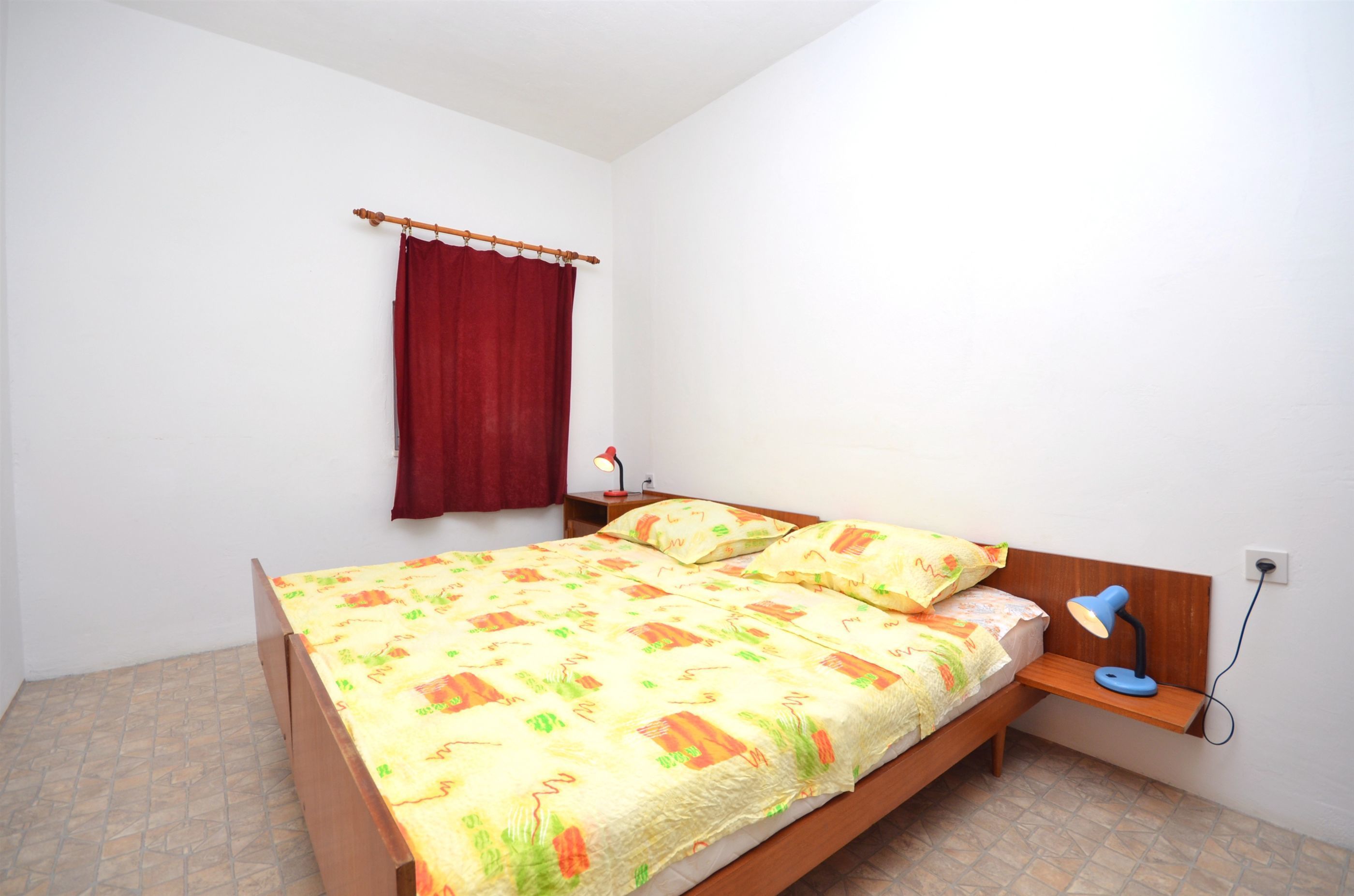 A single bed with a patterned bedspread, a nightstand with a lamp, and a red curtain.