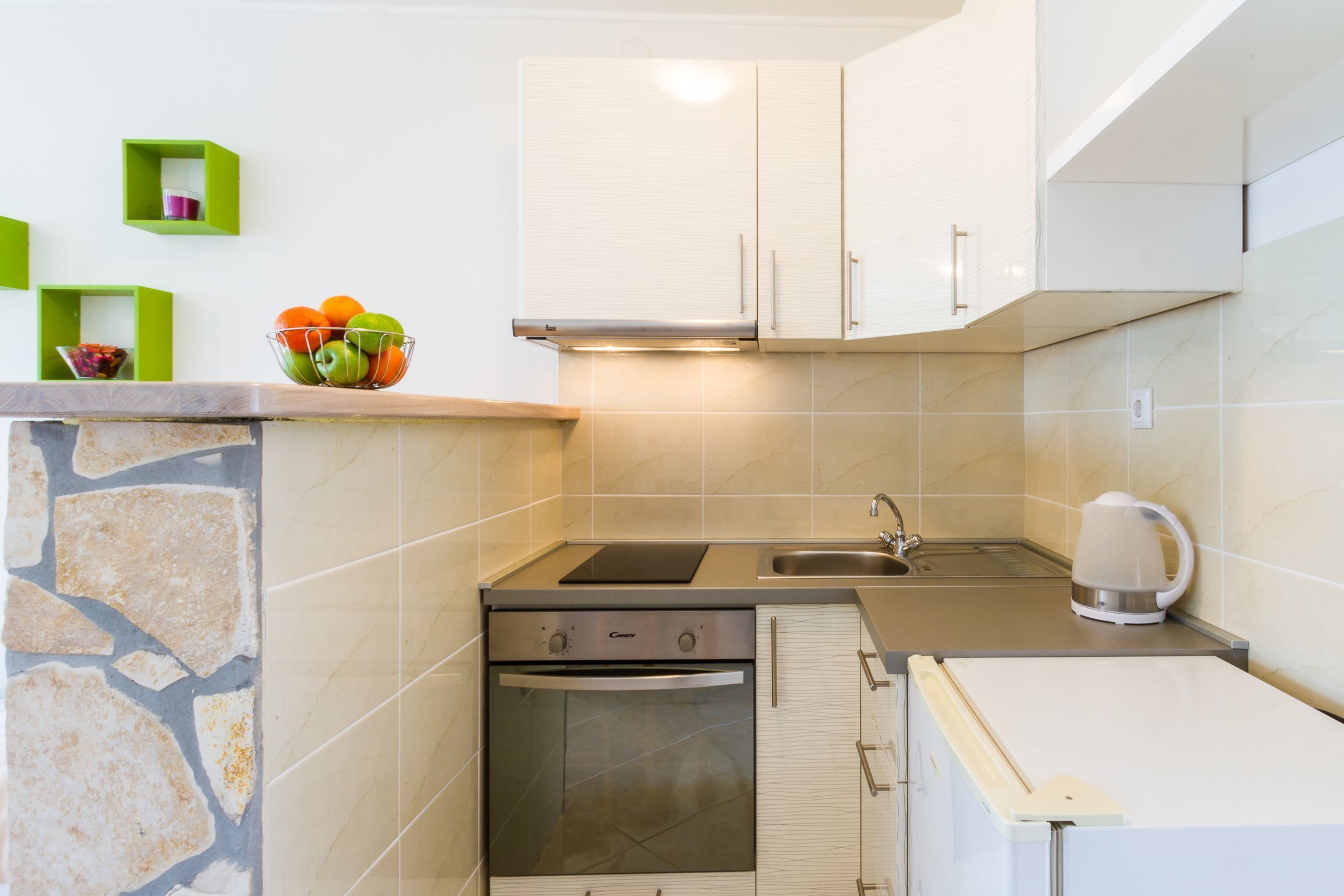 A compact kitchen with a built-in oven, a stovetop, and a sink. There are green storage shelves on the wall and a kettle on the countertop.