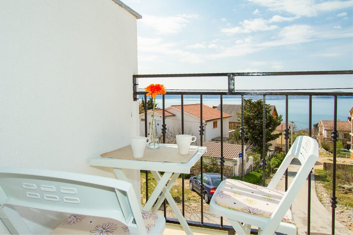 A balcony with a table and chairs, a flower, and a view of houses and the sea.