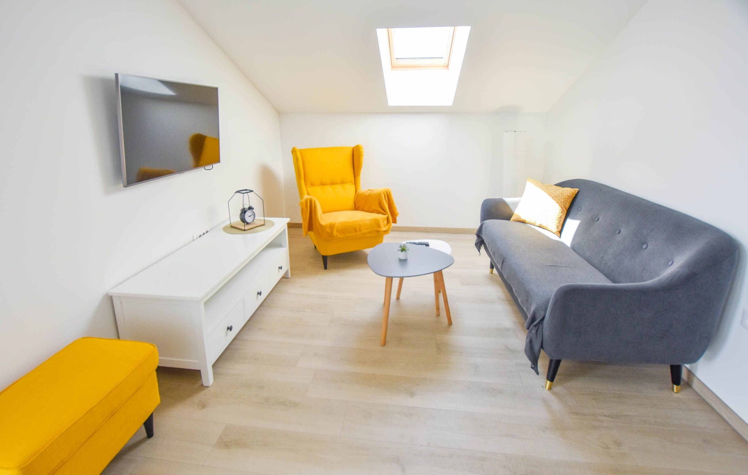 Una sala moderna con una televisión de pantalla plana, un sillón amarillo, un sofá gris, una mesita redonda y un armario blanco.