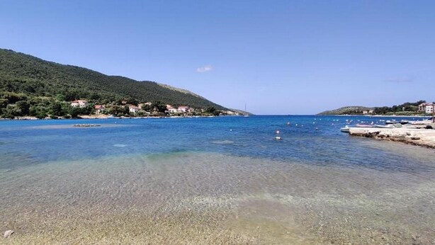 Escena costera con agua cristalina, una playa de guijarros y una colina con edificios.