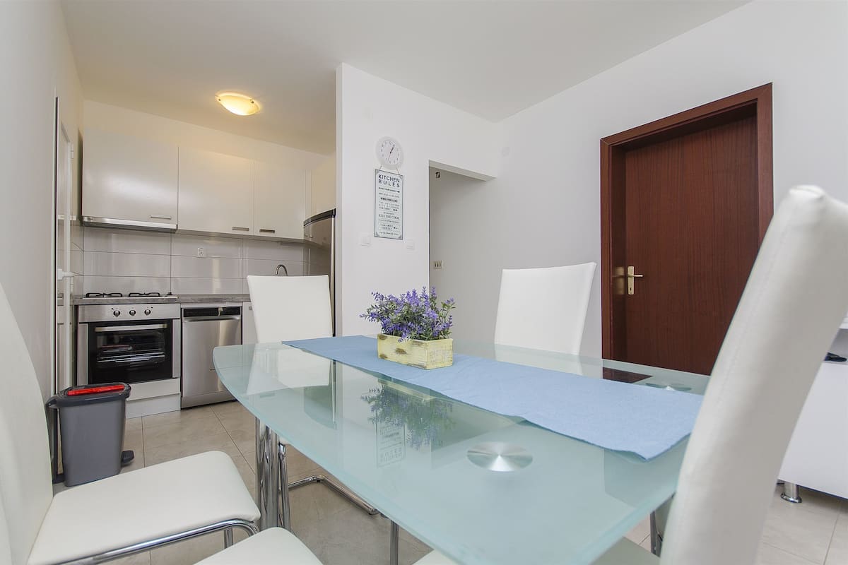 A compact kitchen with stainless steel appliances, a dining table with a glass top, and a wooden door.