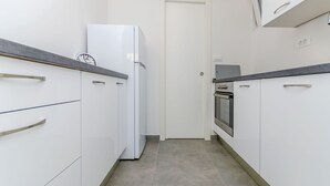 Een smalle keuken met witte kasten, een witte koelkast en een roestvrijstalen oven.