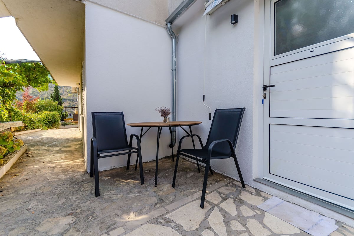 A small patio with a table and two chairs, a white door, and a view of greenery.