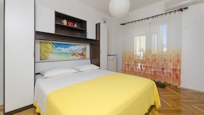 Ein Schlafzimmer mit einem Bett, einem Gemälde, einer Kommode mit Dekorationen, einer weißen Klimaanlage und einem Fenster mit durchsichtigen Vorhängen.