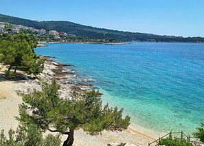 A coastal view with clear blue waters, a sandy beach, and a backdrop of buildings on a hillside.