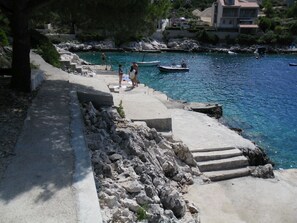 A coastal walkway with steps leading to a rocky shore, boats in the water, and buildings in the background.