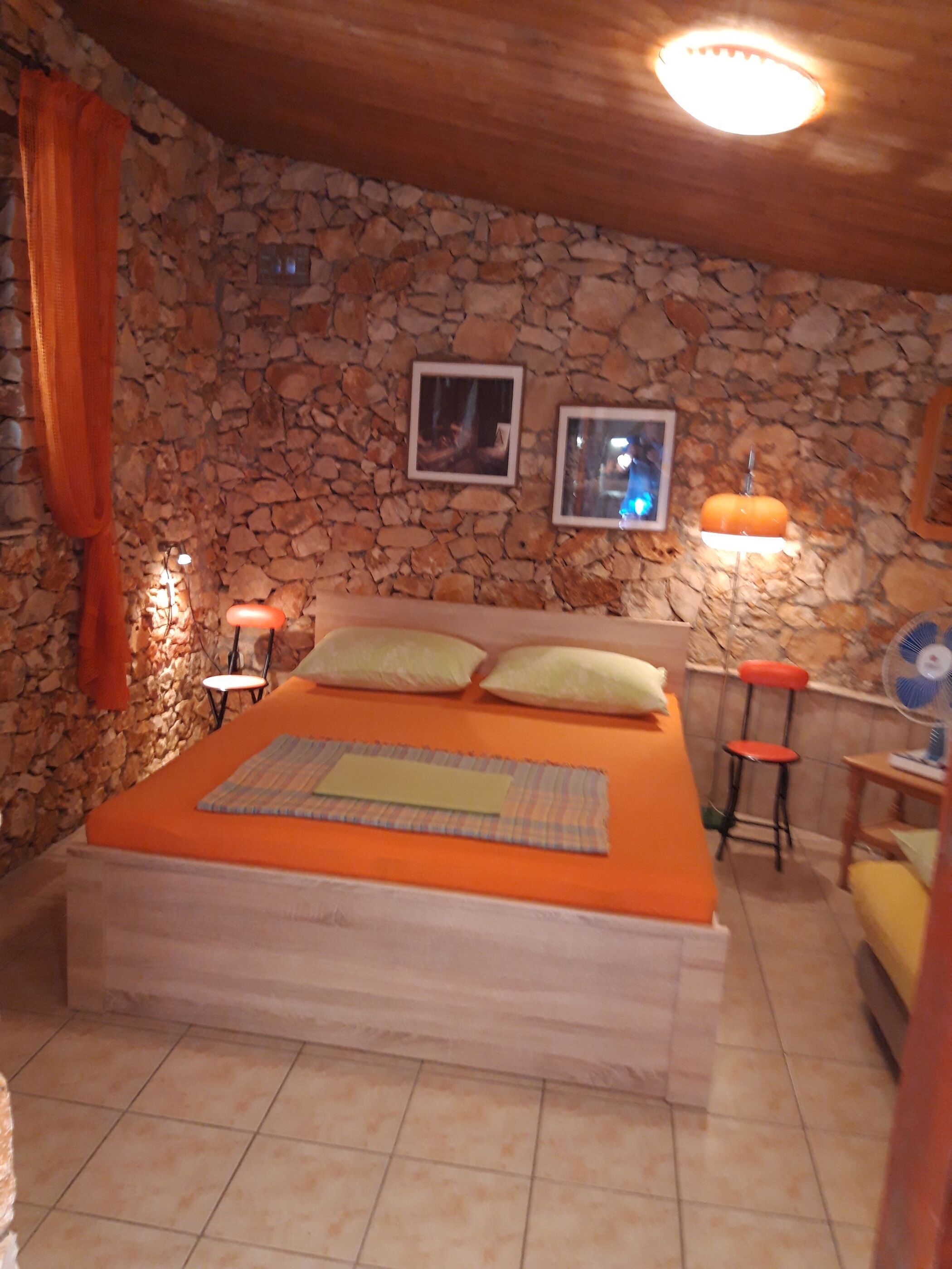 Ein Schlafzimmer mit Steinwand, einem Bett mit orangefarbener Bettdecke, zwei Kissen, einem Nachttisch mit Lampe und einem Sessel.