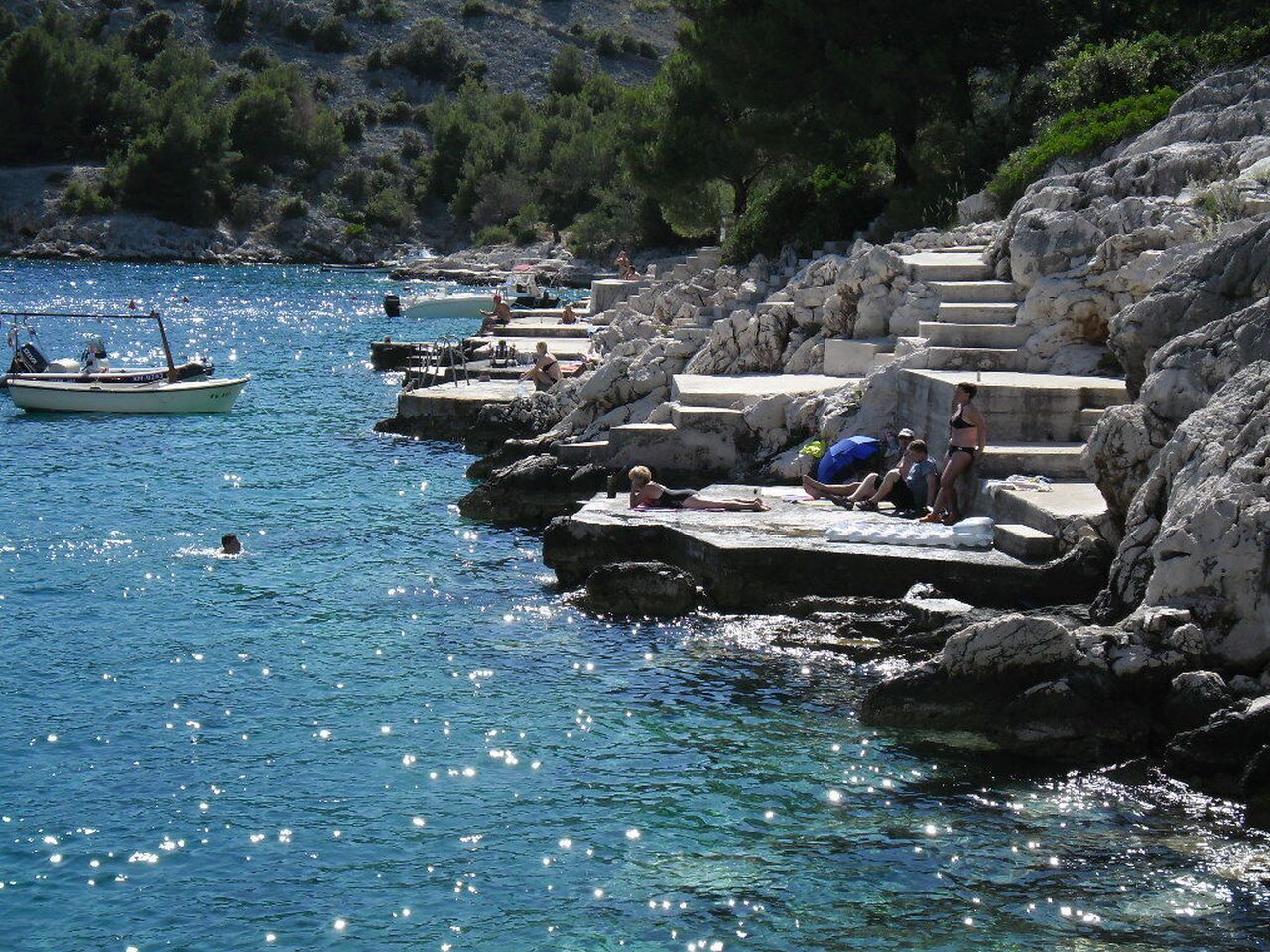 A coastal area with rocky steps leading into the water, a boat on the sea, and people relaxing on the steps.
