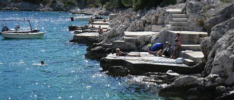 A coastal area with rocky steps leading into the water, a boat on the sea, and people relaxing on the steps.